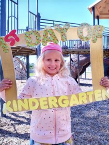 A child holds a handmade paper frame in front of her that reads "1st day of kindergarten."